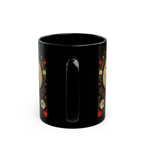 A black ceramic mug featuring a gothic design of a raven against a golden full moon, framed by a floral wreath of coral-colored daisies, white chrysanthemums, and delicate botanical elements. The design includes rich green foliage and small red buds arranged in a symmetrical pattern. The mug has a classic C-shaped handle with a glossy finish.